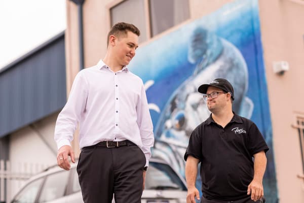 Two people walking and talking outside an industrial building with a large blue mural in the background.