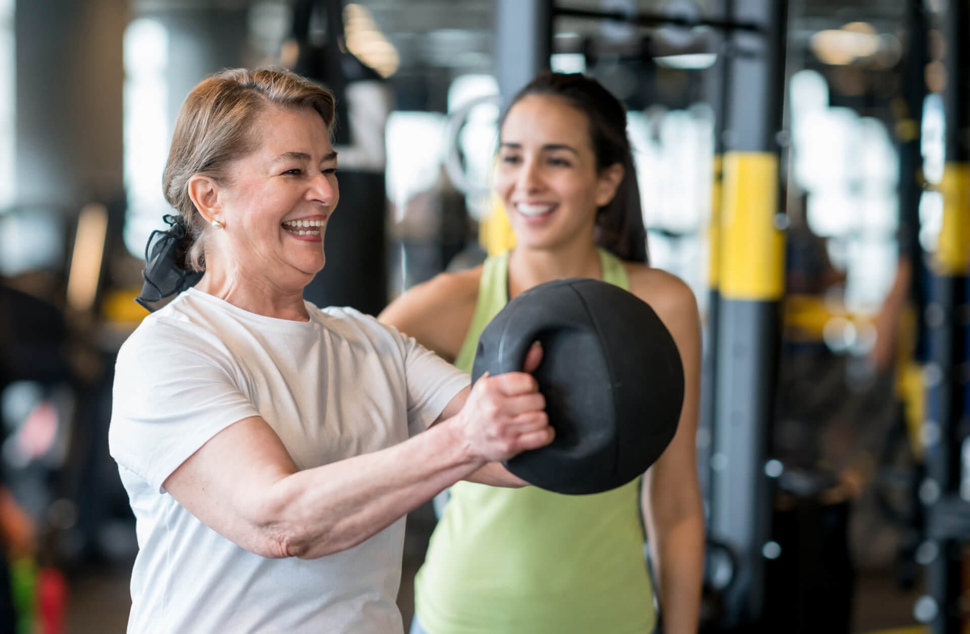 Older woman happily lifting kettlebell with a personal trainer providing encouragement
